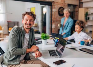 Portrait of a smiling man looking directly into the camera while sitting at a desk and using a laptop to code