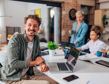 Portrait of a smiling man looking directly into the camera while sitting at a desk and using a laptop to code
